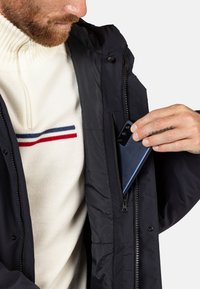 Chaqueta negra con bolsillos con cremallera, llevada sobre un suéter crema con una franja roja y azul. Un teléfono inteligente se está colocando en el bolsillo de la chaqueta.