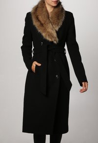 Sort uldfrakke med en brun pelskrave, dobbeltradet design, forlommer og et bånd i taljen. Glat tekstur og skræddersyet pasform.