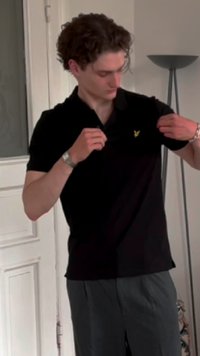 Juoda polo marškinėliai su trumpomis rankovėmis ir maža geltona emblema kairiajame krūtinės srityje, dėvimi su pilkais kelnėmis. Lygi medžiaga, klasikinis dizainas.