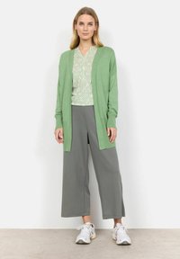 Ljusgrön cardigan med öppen framsida, matchad med en geometriskt mönstrad topp i grön och vit, grå vida byxor och vita sneakers.