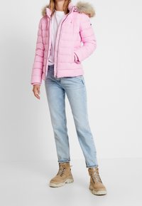 Rosa Daunenjacke mit einer Kapuze mit Futter, horizontalen Nähten und einem Reißverschluss an der Vorderseite. Getragen mit hellblauen Jeans und sandfarbenen Schnürstiefeln.