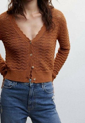 Femme portant un cardigan en maille texturée couleur rouille avec des boutons et un jean bleu taille haute, debout les mains derrière le dos.