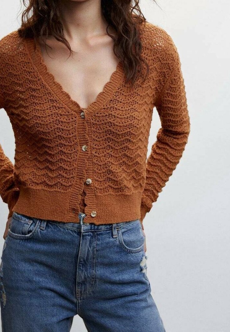 Femme portant un cardigan en maille texturée couleur rouille avec des boutons et un jean bleu taille haute, debout les mains derrière le dos.