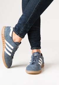 Blue suede athletic shoes with white stripes, rubber outsole, and a textured surface, shown worn with dark blue jeans.