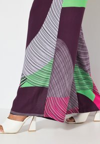 Kleurrijke maxi jurk met geometrische patronen in paars, groen en roze, met een gladde stofafwerking en witte block-heeled sandalen.