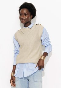 Gilet en tricot beige à motif côtelé, avec des boutons dorés sur les épaules, porté sur une chemise à rayures bleues et blanches, associé à un jean en denim clair.