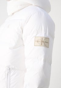 Biała kurtka puchowa z wysokim kołnierzem, zamykaną kieszenią i beżową naszywką z logiem Calvin Klein Jeans na górnej części rękawa.