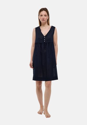 Femme debout pieds nus face à l'avant, portant une robe bleu marine sans manches à col en V et trois boutons sur la poitrine, sur un fond uni.