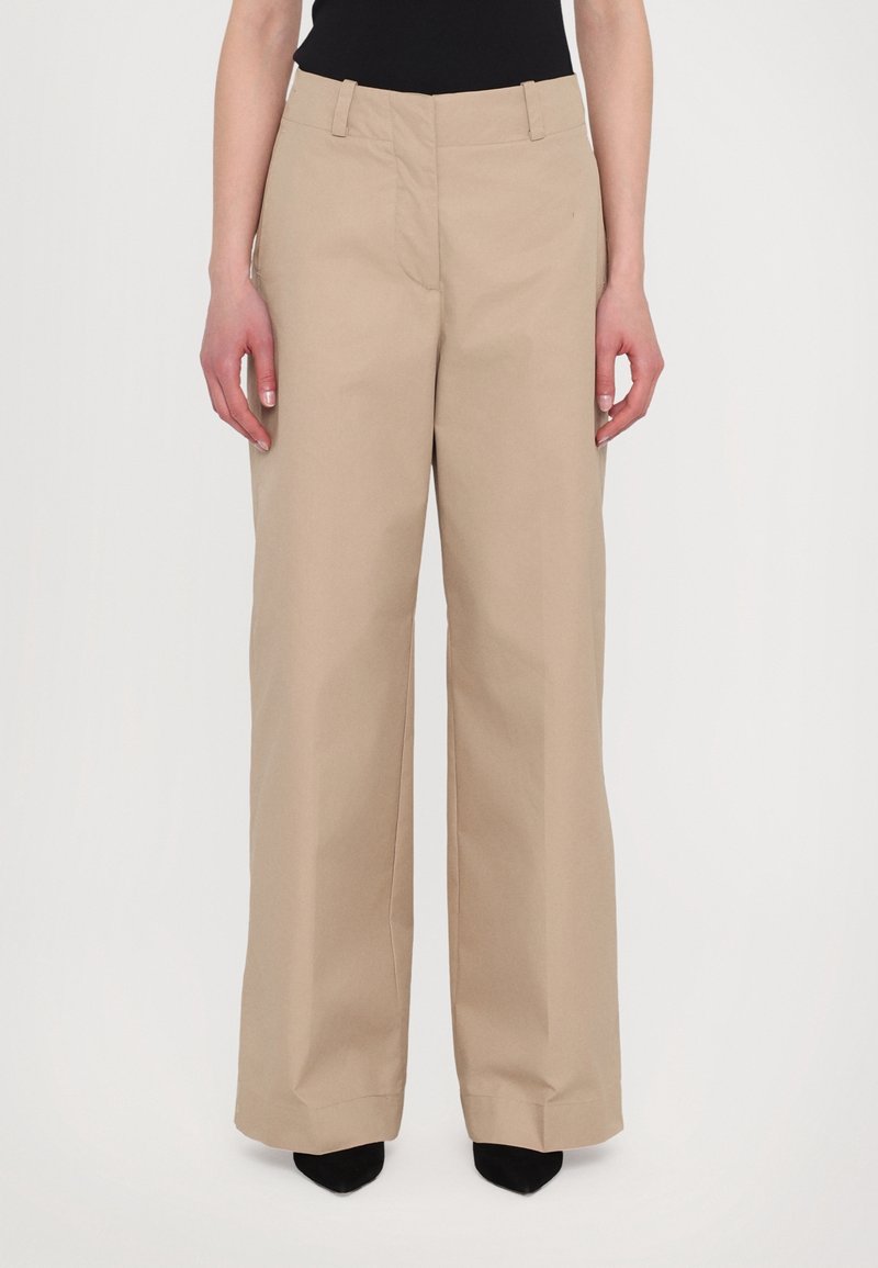 Femme portant un pantalon beige à jambes larges et des chaussures noires à bout pointu, debout devant un fond clair uni.