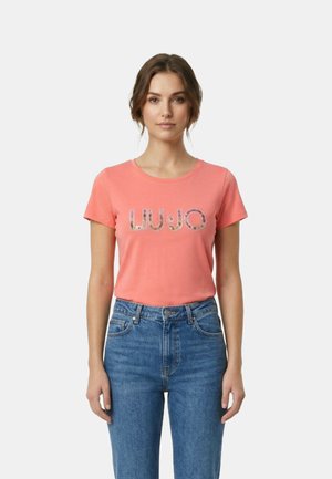 Giovane donna con capelli castani che indossa una t-shirt LUJO corta color corallo e jeans blu a vita alta, in piedi davanti a uno sfondo semplice.