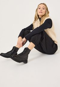 Black leather ankle boots with lace-up design, ribbed texture, and chunky soles. Worn with black pants and a beige knit vest.