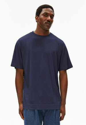 Mann trägt ein schlichtes, dunkelblaues oversized T-Shirt und blaue Jeans, steht vor weißem Hintergrund mit neutralem Gesichtsausdruck.