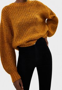 Grober Strickpullover in einem satten Senffarbton, mit rundem Ausschnitt, gerippten Bündchen und lockerem Schnitt. Kombiniert mit schwarzen, eng anliegenden Hosen.