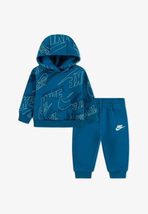 Plava Nike dukserica s prednjim džepom, koja sadrži hrabar print logotipa svuda po površini. Uparena s odgovarajućim trenerkama, oboje od mekanog materijala.