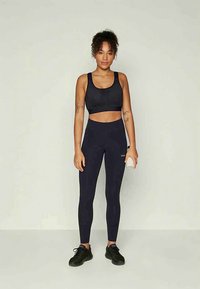 Woman in black sports bra, high-waist leggings, and black sneakers standing against a plain light background holding a small white water bottle.