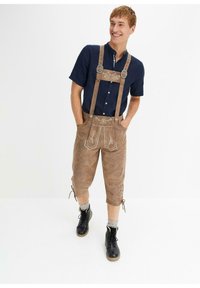 Lederhosen in suede marrone con dettagli ricamati, bretelle regolabili e bottoni; abbinate a una camicia blu navy a maniche corte e stivali neri.