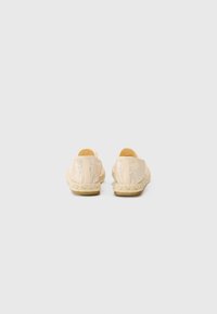 Strukturert beige slip-on sko med et vevd design, elastiske detaljer og en naturlig tau-såle, sett bakfra.
