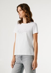 Mladá žena s vlnitými hnědými vlasy má na sobě bílý tričko s ozdobným logem a světle šedé džíny, stojí na pozadí na čistě bílém pozadí.