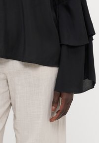 Black, layered blouse with flared sleeves; paired with light, textured beige trousers. Smooth fabric with a slight sheen.
