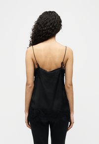 Γυναίκα με μακριά σγουρά μαλλιά που φοράει ένα μαύρο καλοκαιρινό τοπ με λεπτές τιράντες και δαντέλα, από πίσω, με φόντο απλή επιφάνεια.