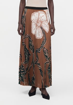 Femme portant une jupe maxi plissée marron avec de grands motifs floraux blancs et noirs, associée à des talons noirs pointus.