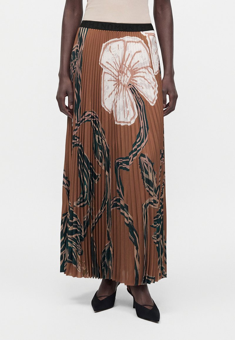Femme portant une jupe maxi plissée marron avec de grands motifs floraux blancs et noirs, associée à des talons noirs pointus.