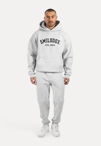 Grau Kapuzenpullover mit schwarzem Aufdruck "SMILODOX ATHL. WEAR", kombiniert mit passender grauer Jogginghose und weißen Sneakern, alles in einem lässigen Fit.