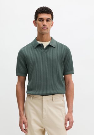 Mężczyzna ubrany w zieloną, prążkowaną koszulkę polo z krótkim rękawem, na beżową podkoszulkę, w beżowych spodniach, stojący na tle białej ściany.