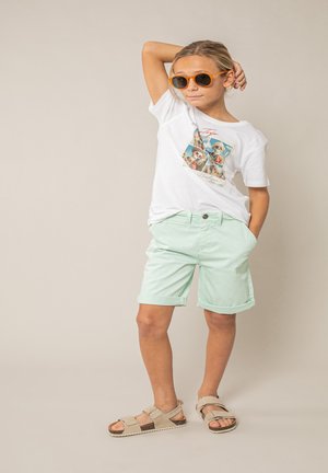 Enfant portant des lunettes de soleil orange, un t-shirt blanc avec un motif graphique, un short vert menthe et des sandales beige, se tenant debout avec une main derrière la tête.