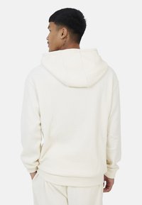 Hoodie color crema realizzato in tessuto a coste, presenta un cappuccio con cordino e maniche lunghe con orli a costine. Dettagli di texture sottili visibili.