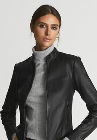 Černá kožená bunda s hladkým povrchem, na zip vpředu a bez límce. Nosená přes šedý žebrovaný rolák. Obsahuje boční kapsu na zip.