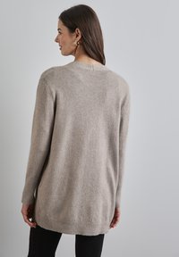 Pull en tricot beige avec une coupe décontractée, des poignets et un ourlet côtelés, et un détail de couture dans le dos. Le tissu semble doux etTexturé.