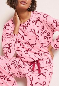 Rosa pyjamaset med långärmade tröjor och byxor som har ett upprepat mönster av mörkröda rosetter och hjärtan. Mjuk tyg med avslappnad passform och dragsko.