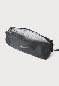 Zwarte nylon etui met een grijze voering, met ritssluiting en een klein handvat. Voorzien van een wit Nike-logo aan de voorkant. Compacte en rechthoekige vorm.