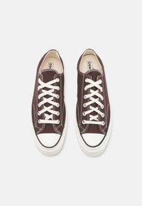 Braune Canvas-Sneaker mit weißen Schnürsenkeln, Gummizahnkappe und kontrastierenden Nähten. Flaches Design, niedriges Profil und strukturierte Oberfläche.