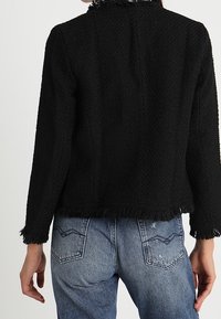 Veste noire tissée avec une finition texturée, présentant des bords effilochés. Associée à un jean en denim bleu clair avec des poches arrière courbées.