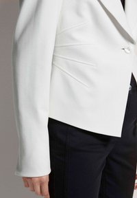 Weiße taillierte Blazerjacke mit strukturierten Nähten und glatter Textur, mit einem einzelnen Knopfverschluss und einer maßgeschneiderten Silhouette, kombiniert mit dunklen Hosen.