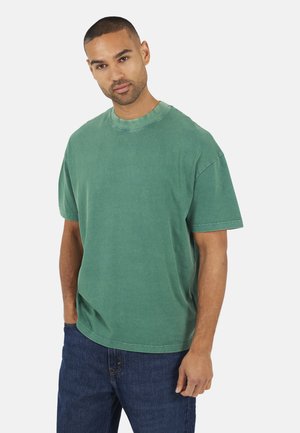 Uomo con capelli corti che indossa una maglietta verde larga e jeans blu, in piedi con una mano in tasca contro uno sfondo semplice.