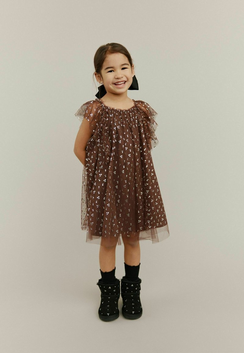 Hnedé tylové šaty s volánovými rukávmi a striebornými hviezdami, skombinované s čiernymi členkovými topánkami a ponožkami po kolená.