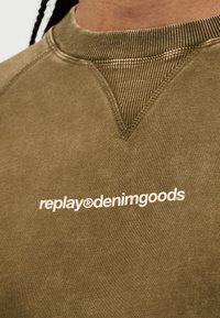 Gros plan d'un sweat-shirt vert olive avec détail de surjet côtelé en V et inscription blanche "replay®denimgoods" imprimée sur la poitrine.
