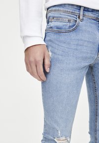 Jeans de mezclilla azul claro con un leve desgastado, diseño de cinco bolsillos y acentos de costura. La tela tiene una apariencia suave y ligeramente texturizada.
