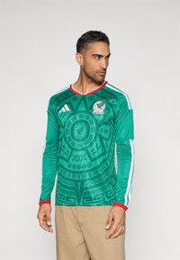 Man i grön Mexiko-fotbollströja med aztekiskalenderdesign, röd krage och manschetter, samt beiga byxor, som tittar åt sidan.