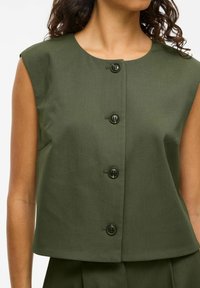 Kurzärmeliges grünes Cropped-Top aus strukturiertem Stoff, mit fünf schwarzen Knöpfen vorne und einem runden Ausschnitt.