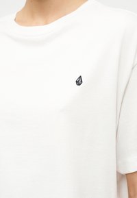 Άσπρο μπλουζάκι με στρογγυλή λαιμόκοψη, με ένα μικρό μαύρο γεωμετρικό λογότυπο κεντημένο στο αριστερό στήθος, που φοριέται από άτομο ορατό από τον λαιμό έως τη μέση.