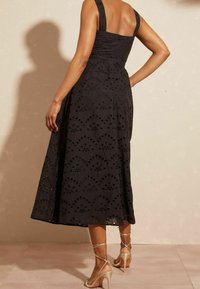 Žena v černých bezrukávových midi šatech s očkovými vzory, s béžovými páskovými lodičkami na vysokém podpatku, stojící s jednou rukou na bocích proti jednoduché zdi.