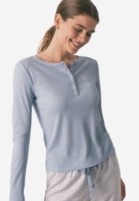 Kvinde if�ørt en lyseblå langærmet henley skjorte og prikkede pajama bukser med snor, smilende med lukkede øjne mod en ensfarvet baggrund.