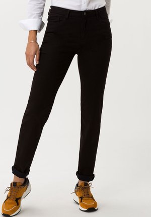 Person wearing black slim-fit pants rolled at the ankles, white shirt with sleeves rolled, and orange sneakers with black details.