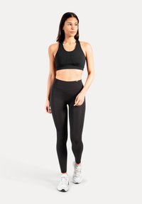 Zwarte atletische sportbeha en hooggetailleerde leggings, beide in effen zwart met een gladde textuur. Model draagt witte sneakers.