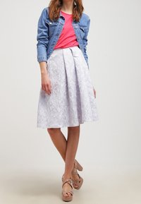 Donna che indossa una giacca di jeans blu sopra una maglietta rosa brillante, una gonna a pieghe bianca con fiori e sandali con piattaforma beige, in piedi contro uno sfondo neutro.