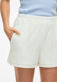 Žena v texturovaných bílých šortkách s elastickým pasem a bočními kapsami, s rukou položenou v levé kapse, v kombinaci se světle modrým topem.